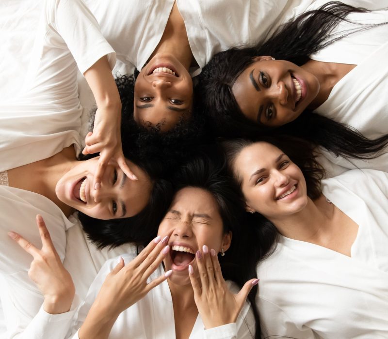 Five happy pretty diverse young girls friends wear robes lying in circle on bed laughing having fun, smiling multi ethnic ladies looking at camera celebrate hen bachelorette party, top view portrait